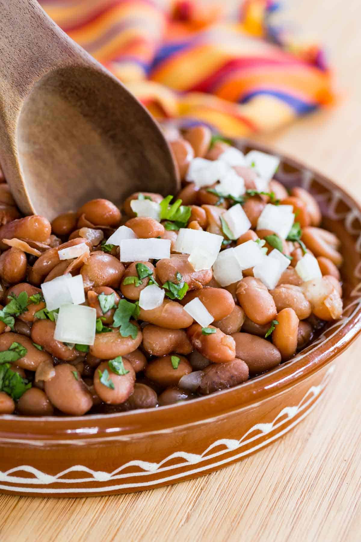 A wooden spoon dips into a bowl of Instant Pot pinto beans garnished with diced onions and cilantro.