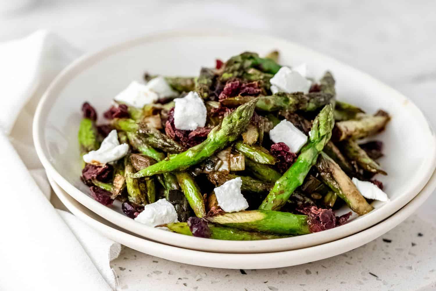 Roasted asparagus salad topped with chunks of goat cheese and dried cherries in two stacked white bowls.