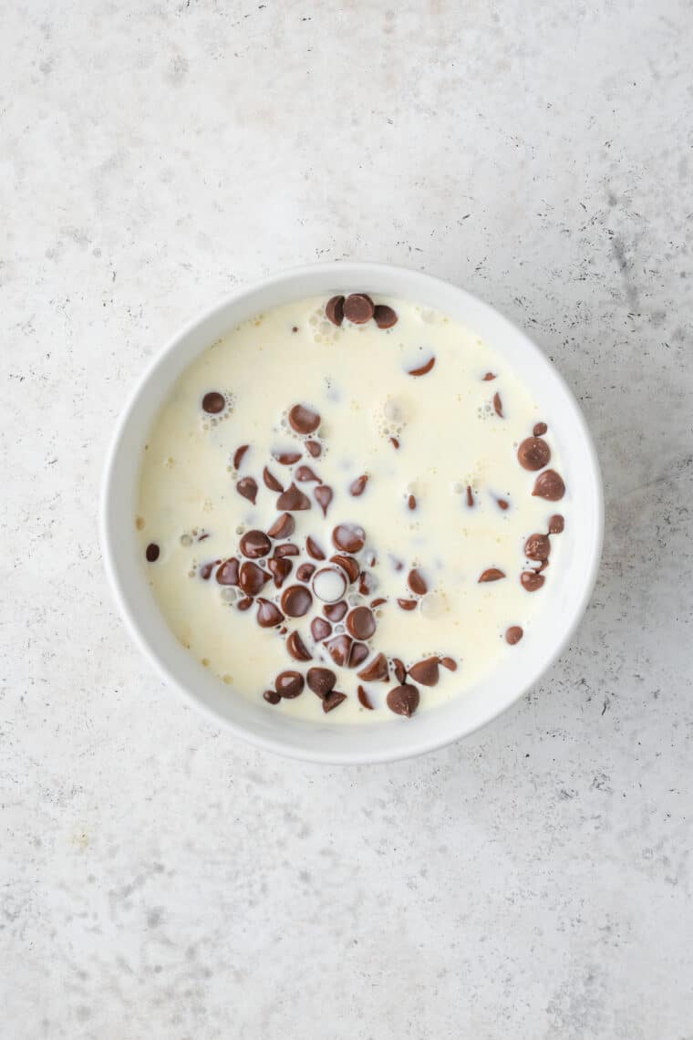 Chocolate chips combined with hot heavy cream inside a white bowl.