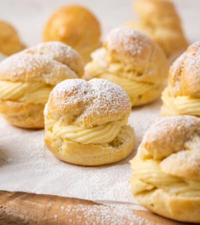 Assorted gluten-free cream puffs filled with vanilla pastry cream and dusted with powdered sugar on a parchment-lined wooden board.