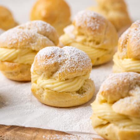 Assorted gluten-free cream puffs filled with vanilla pastry cream and dusted with powdered sugar on a parchment-lined wooden board.