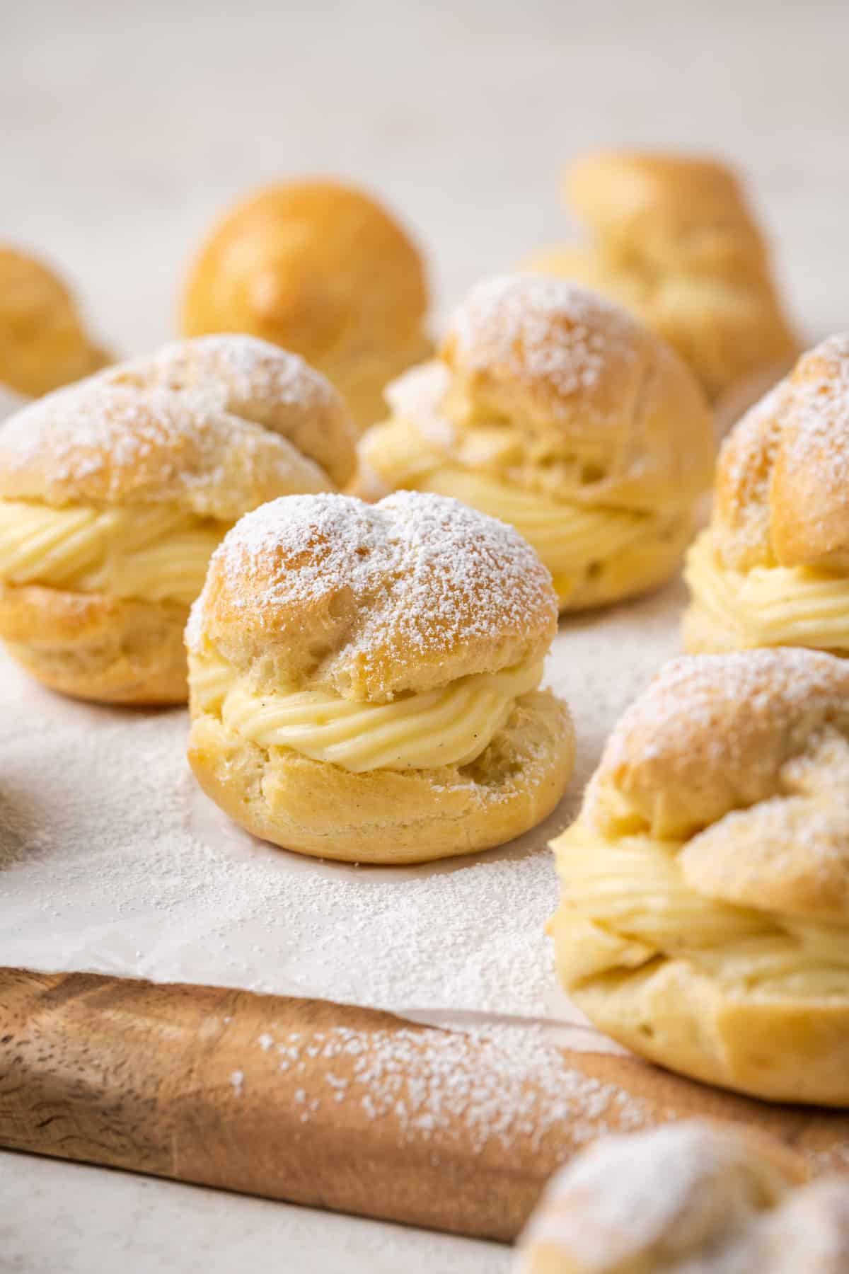 Assorted gluten-free cream puffs filled with vanilla pastry cream and dusted with powdered sugar on a parchment-lined wooden board.