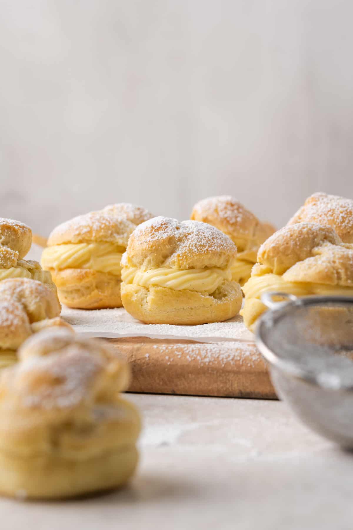 Assorted gluten-free cream puffs filled with vanilla pastry cream and dusted with powdered sugar on a parchment-lined wooden board, with a mesh sieve in the foreground.