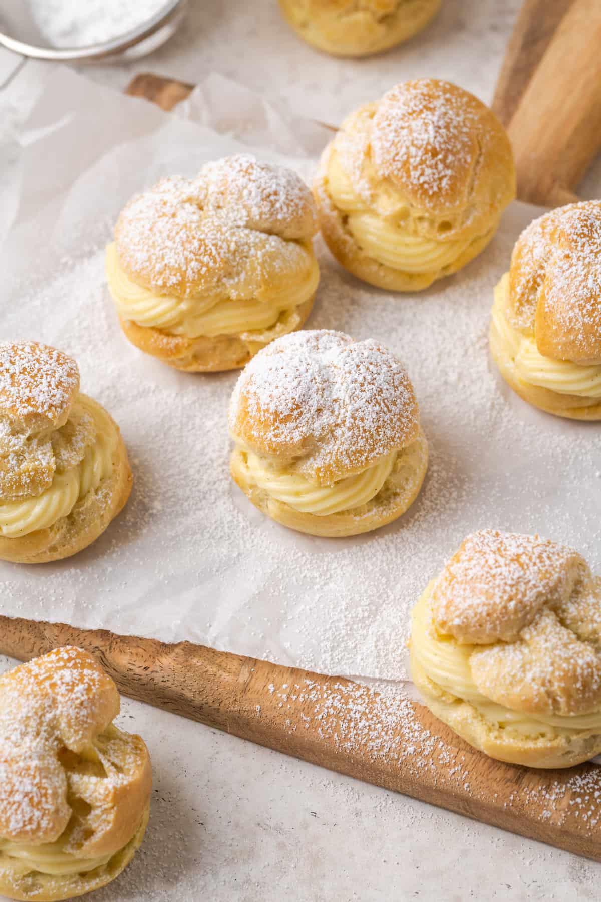 Overhead view of gluten-free cream puffs filled with vanilla pastry cream and dusted with powdered sugar on a parchment-lined wooden board.