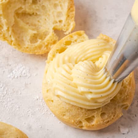 A piping tip pipes vanilla pastry cream into one half of a gluten-free cream puff.