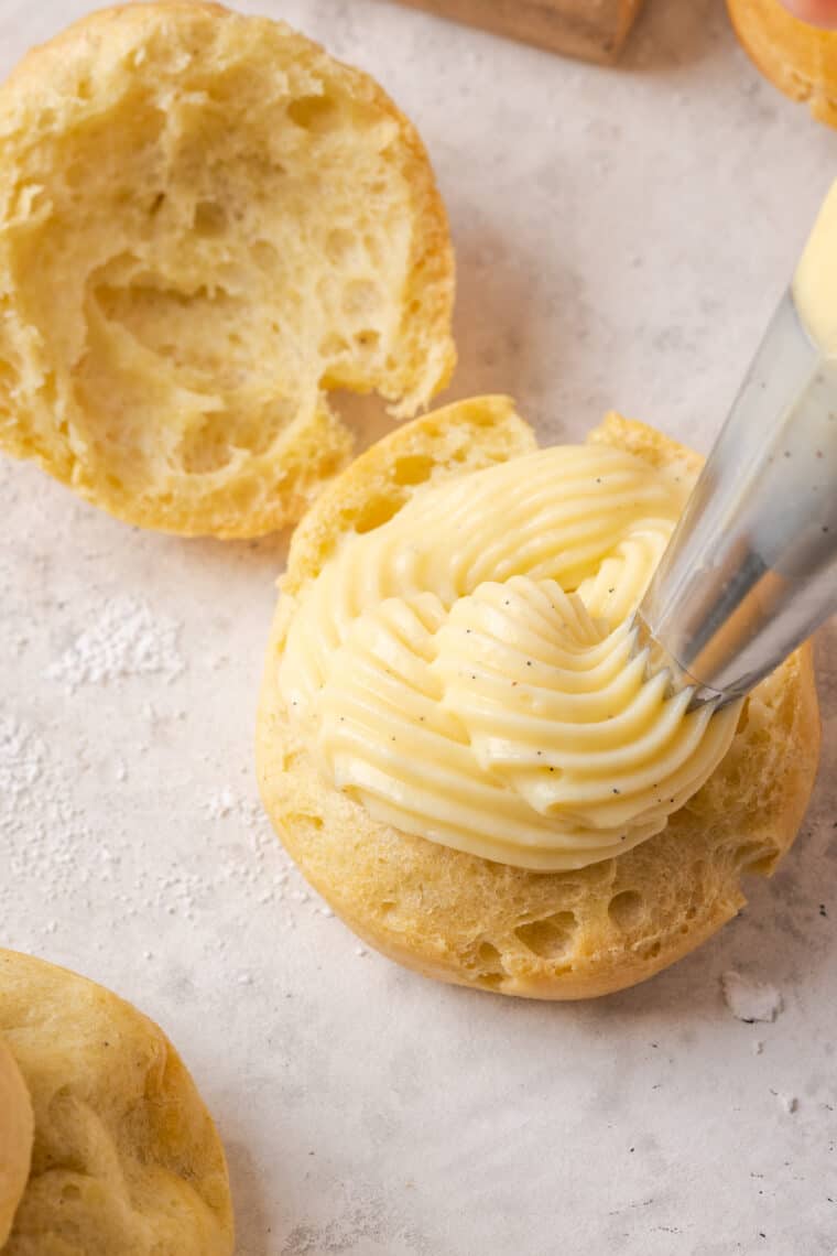 A piping tip pipes vanilla pastry cream into one half of a gluten-free cream puff.