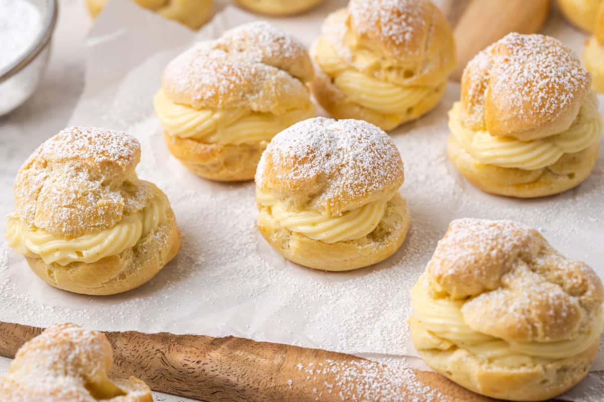 Assorted gluten-free cream puffs filled with vanilla pastry cream and dusted with powdered sugar on a parchment-lined wooden board.