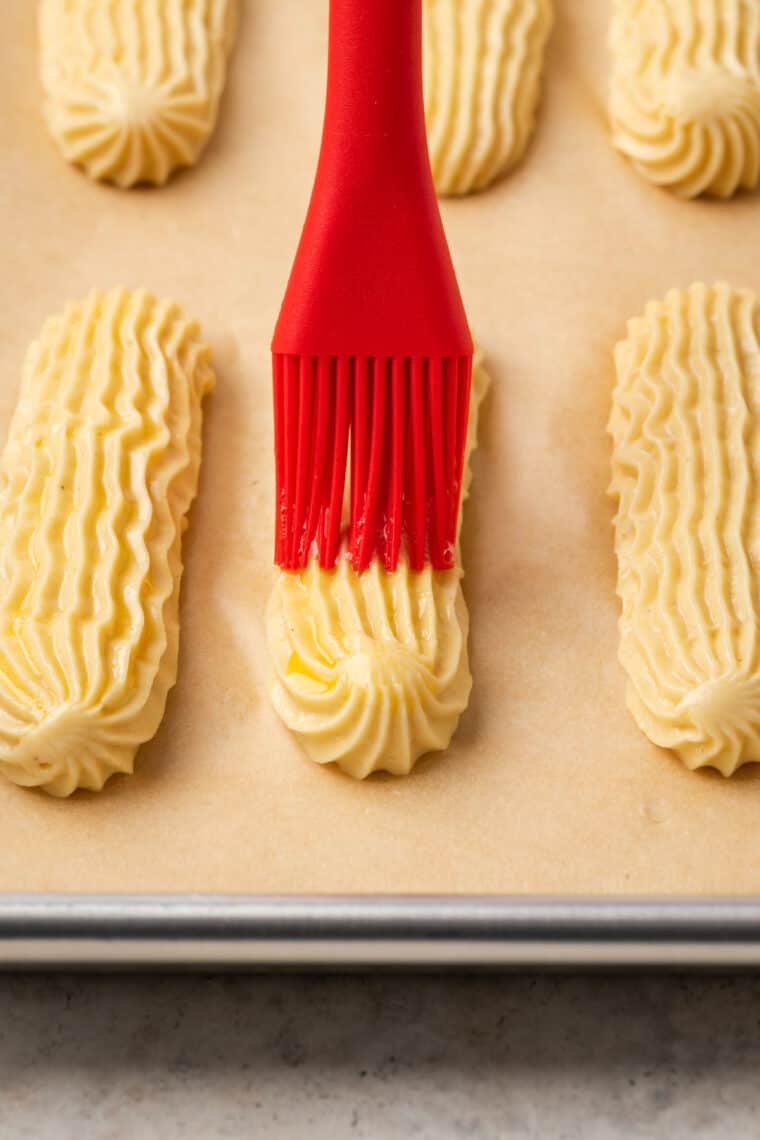 A red rubber brush is used to brush egg wash over piped logs of pate a choux on a parchment-lined baking sheet.