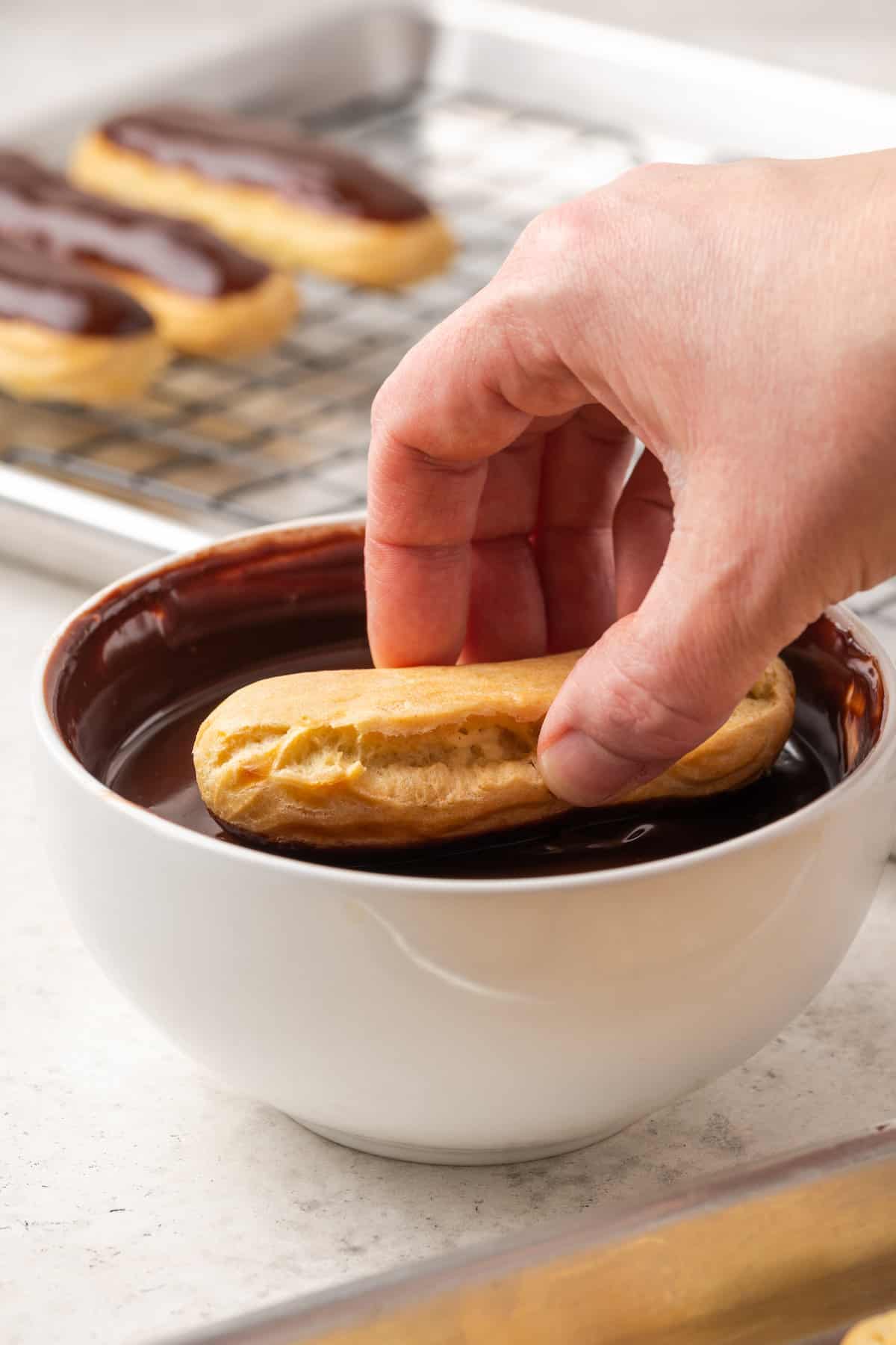 A hand dips a gluten-free eclair into a white bowl of chocolate glaze.