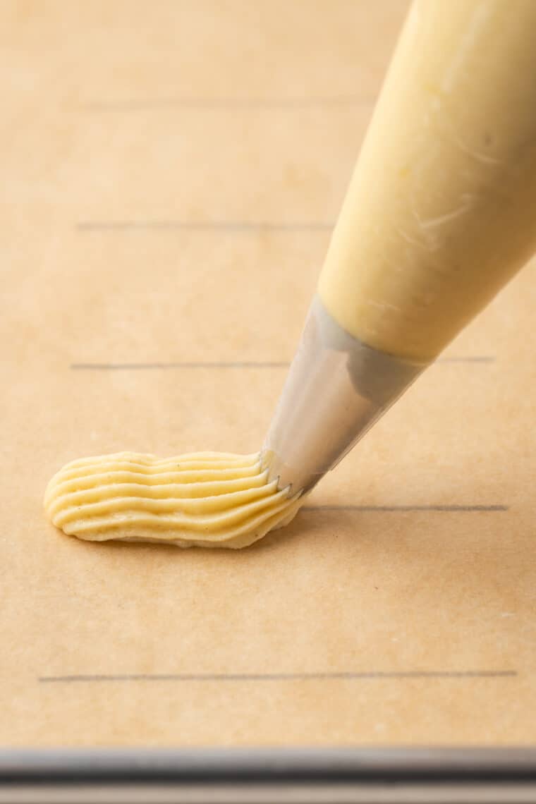 Gluten-free choux pastry is piped from a piping bag onto a parchment-lined baking sheet.
