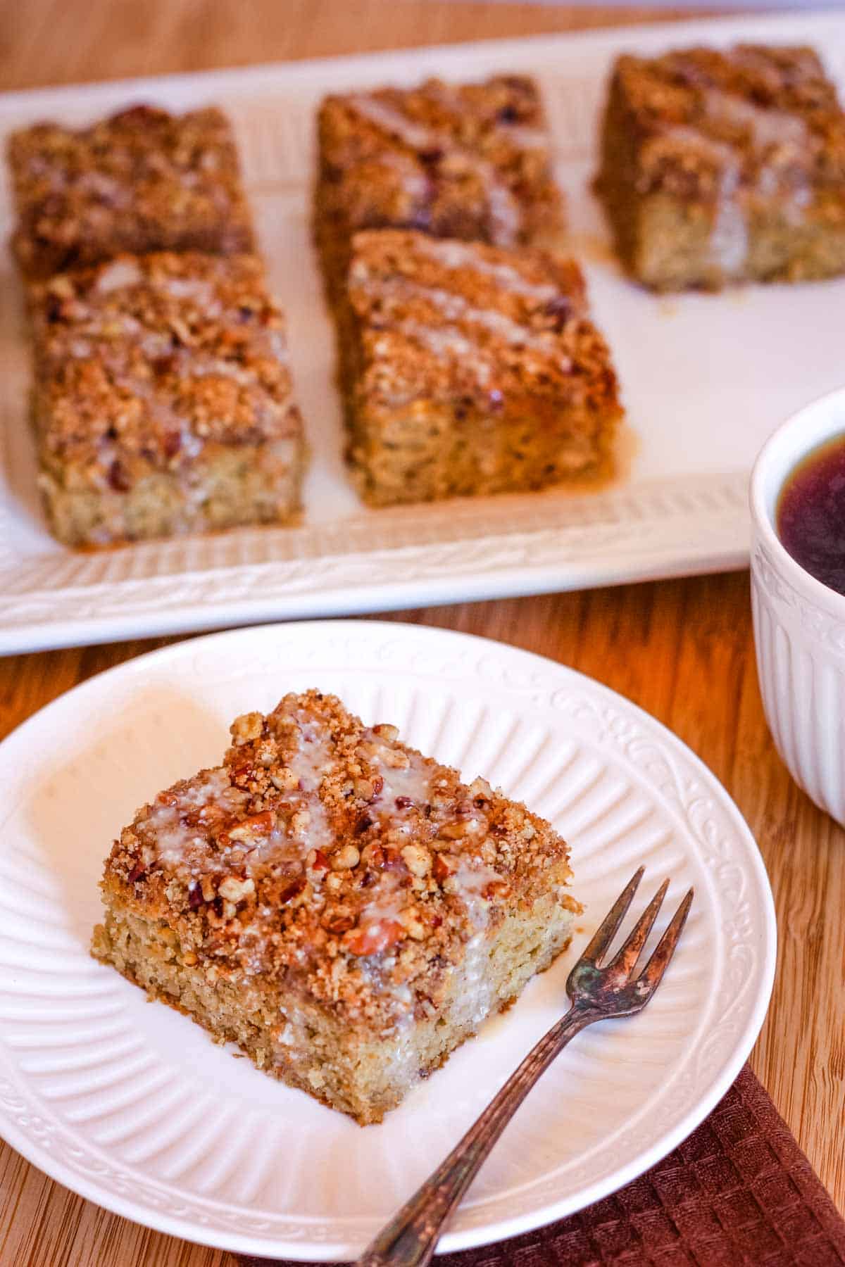 A slice of pumpkin coffee cake on a white plate with a fork, with more slices on a platter in the background.