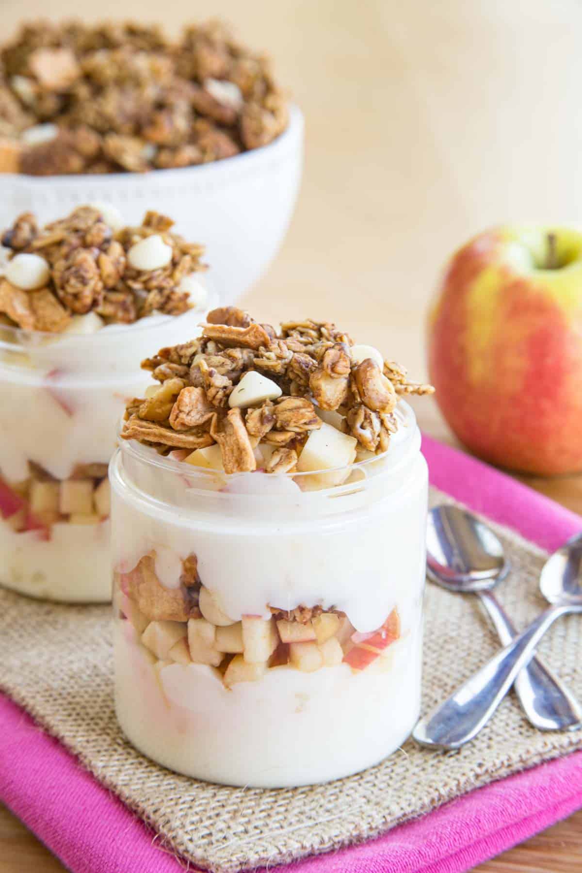 Two apple parfaits on a piece of burlap and pink cloth napkin with a bowl of granola behind them.