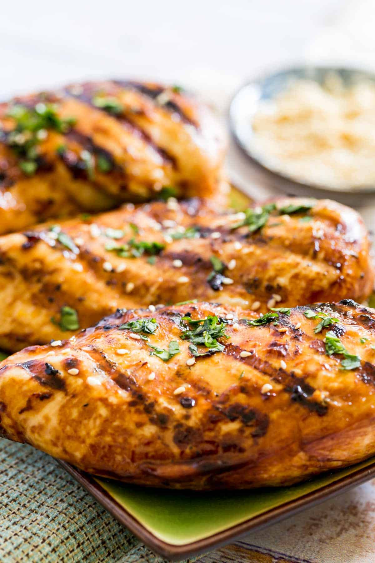 Asian grilled chicken breasts on a rectangular plate garnished with chopped cilantro and sesame seeds.