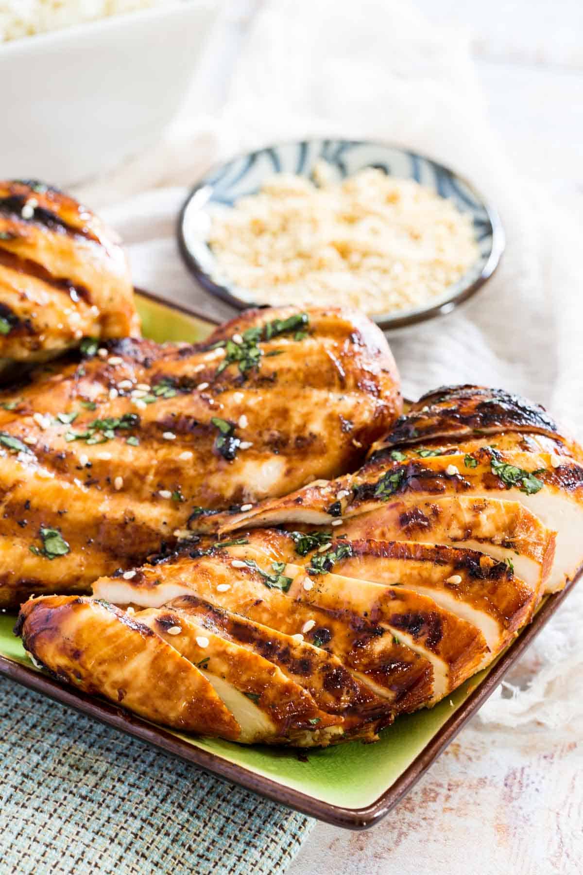 Sliced Asian grilled chicken breasts on a rectangular plate garnished with chopped cilantro and sesame seeds.