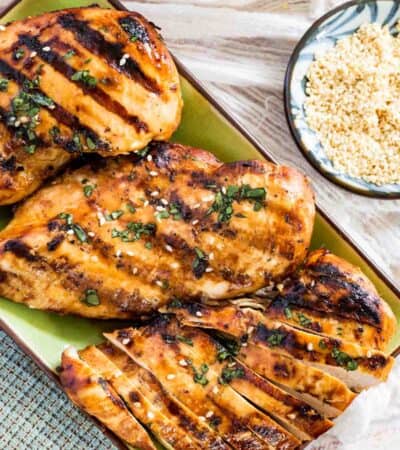 Overhead view of Asian grilled chicken breasts on a rectangular plate garnished with chopped cilantro and sesame seeds.
