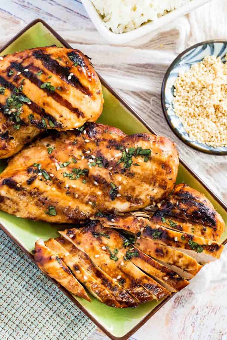 Overhead view of Asian grilled chicken breasts on a rectangular plate garnished with chopped cilantro and sesame seeds.