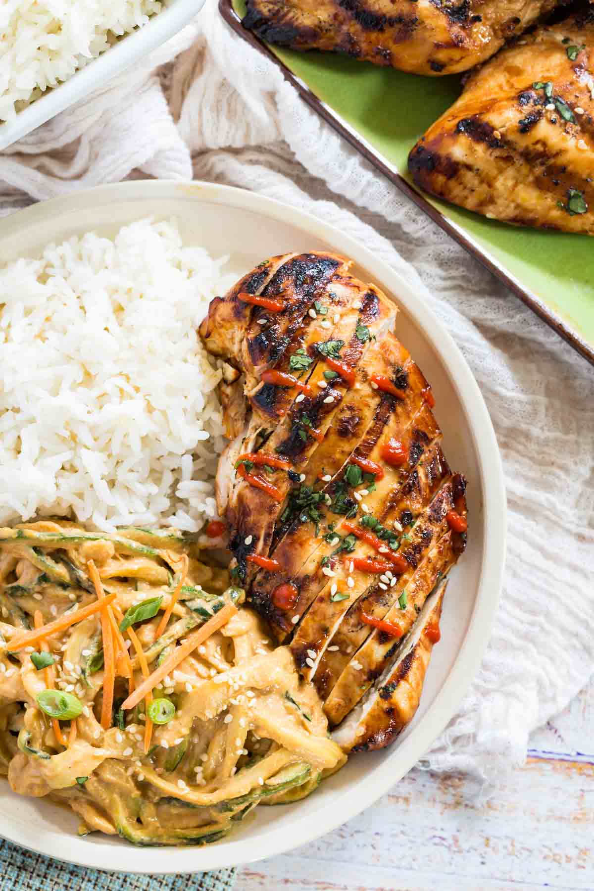 Overhead view of Asian grilled chicken sliced on a plate next to spicy sesame noodles and white rice.