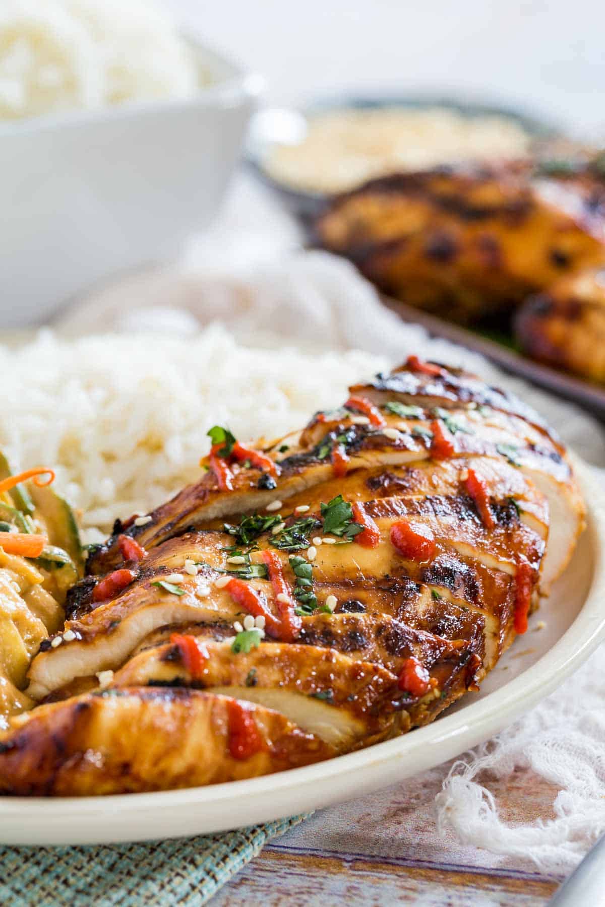 Asian grilled chicken sliced on a plate next to spicy sesame noodles and white rice.