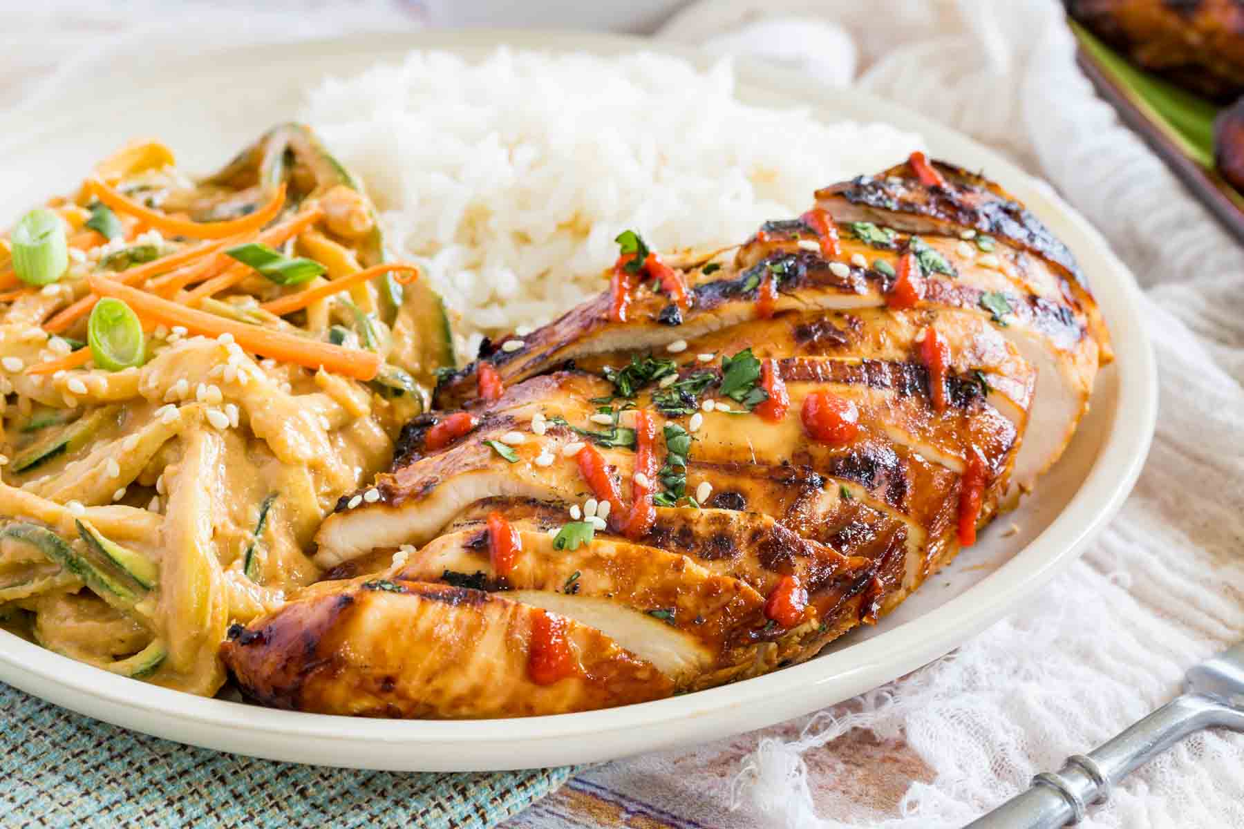 Asian grilled chicken sliced on a plate next to spicy sesame noodles and white rice.
