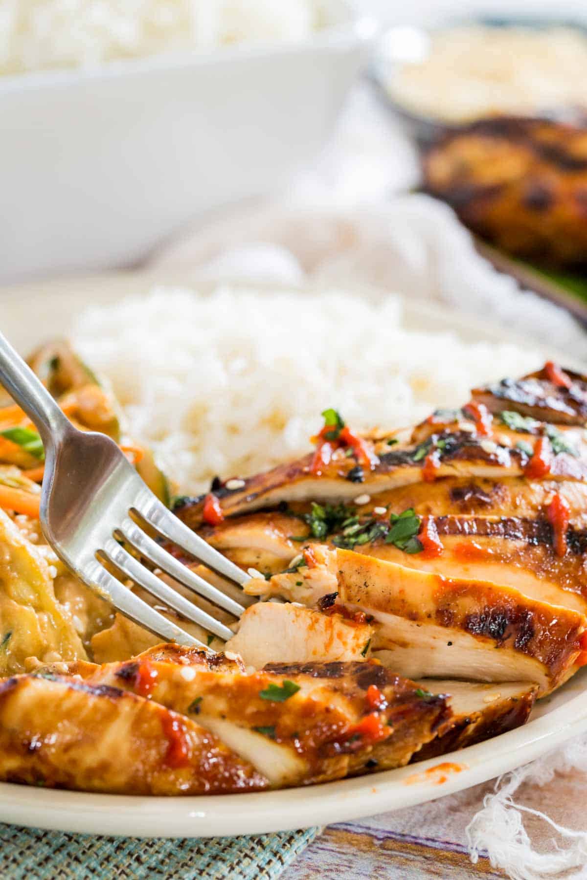 A fork stuck into a slice of Asian grilled chicken on a plate next to spicy sesame noodles and white rice.