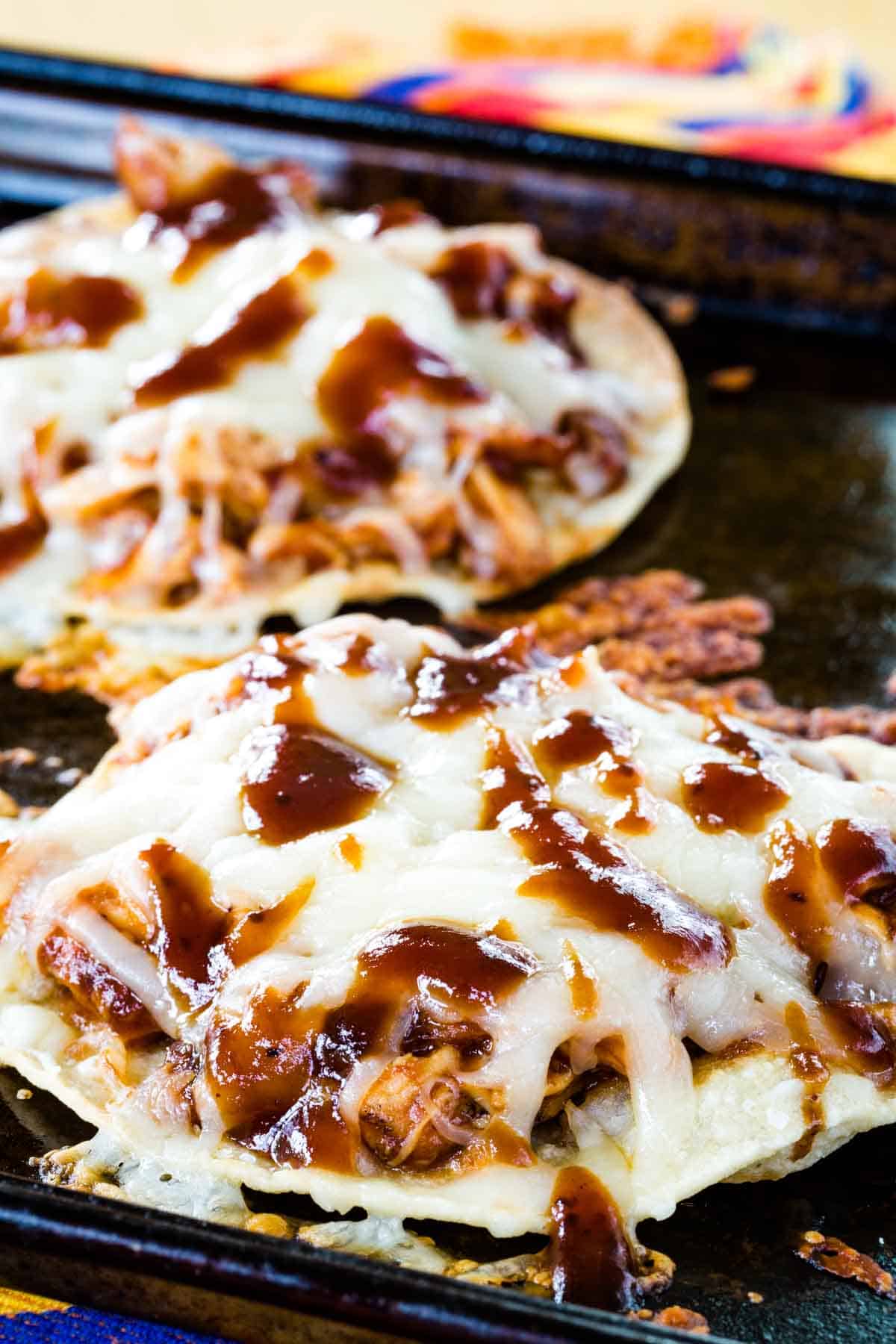 Two BBQ Chicken Tostadas drizzled with barbecue sauce on a baking sheet.