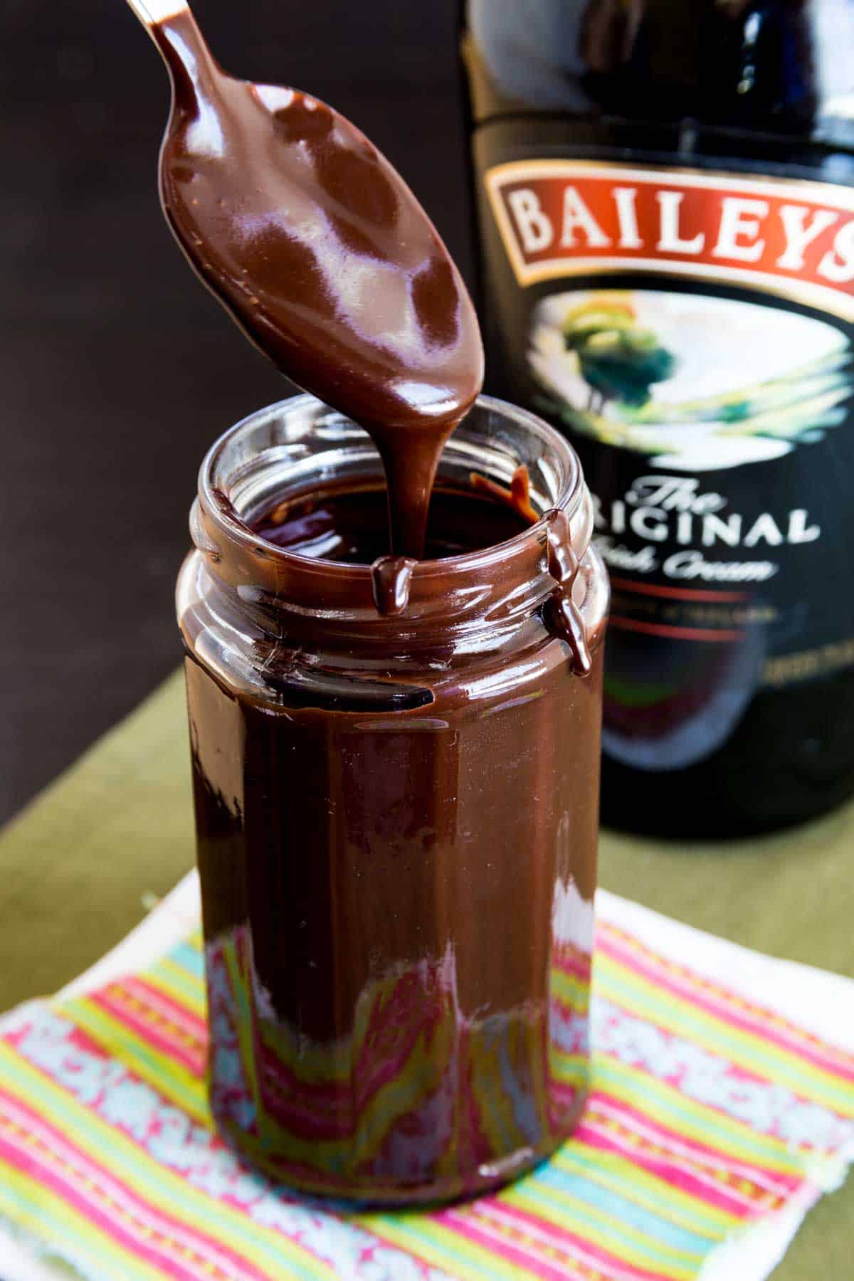 Thick chocolate sauc dripping off a spoon into a jar with a bottle of Bailey's Irish Cream in the background.