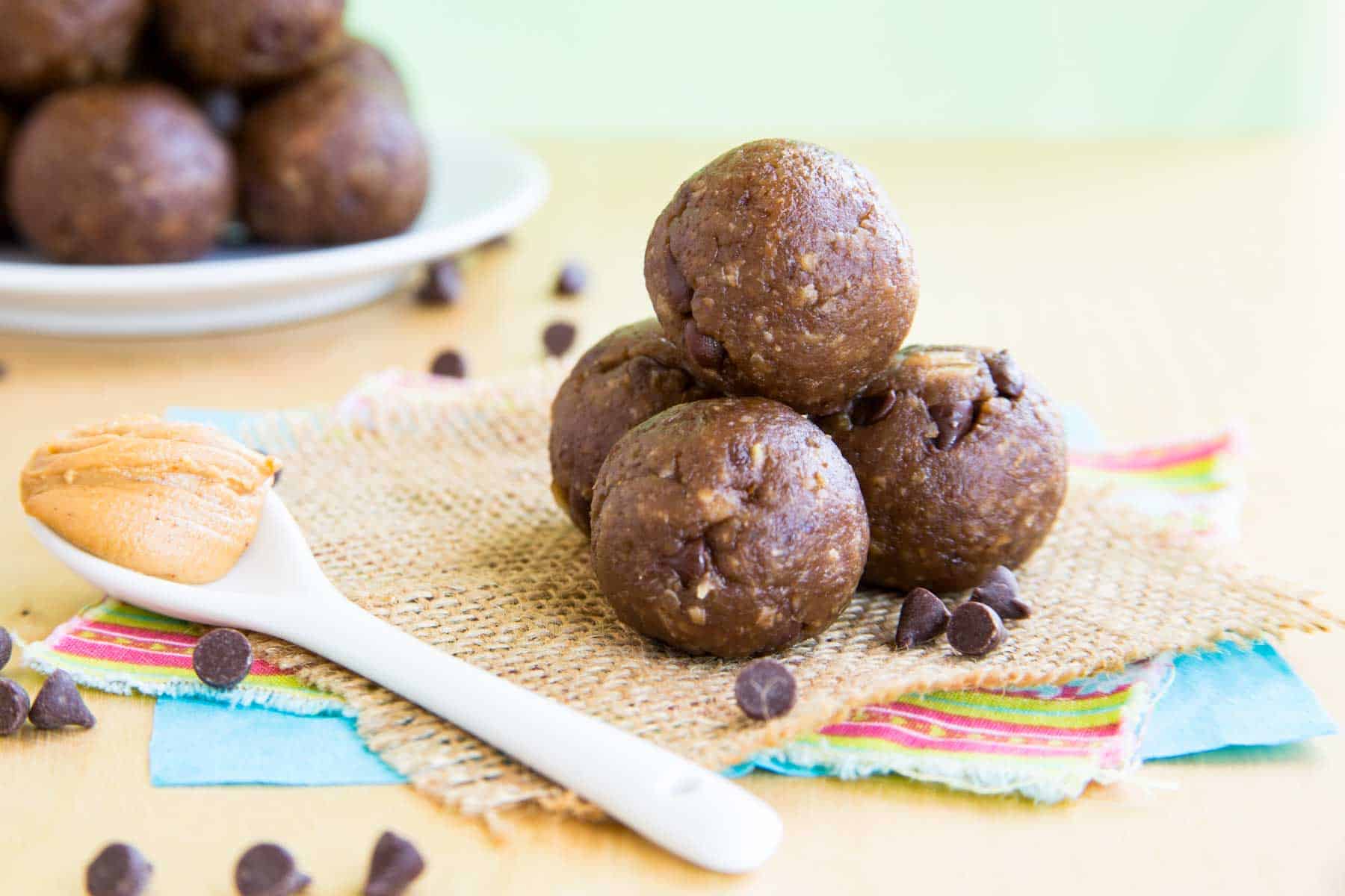 Four energy balls in a pyramid on a piece of burlap and some fabric surrounded by mini chocolate chips and white spoon with peanut butter.