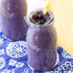 A blueberry smoothie in a glass bottle with four small blueberries floating on top.
