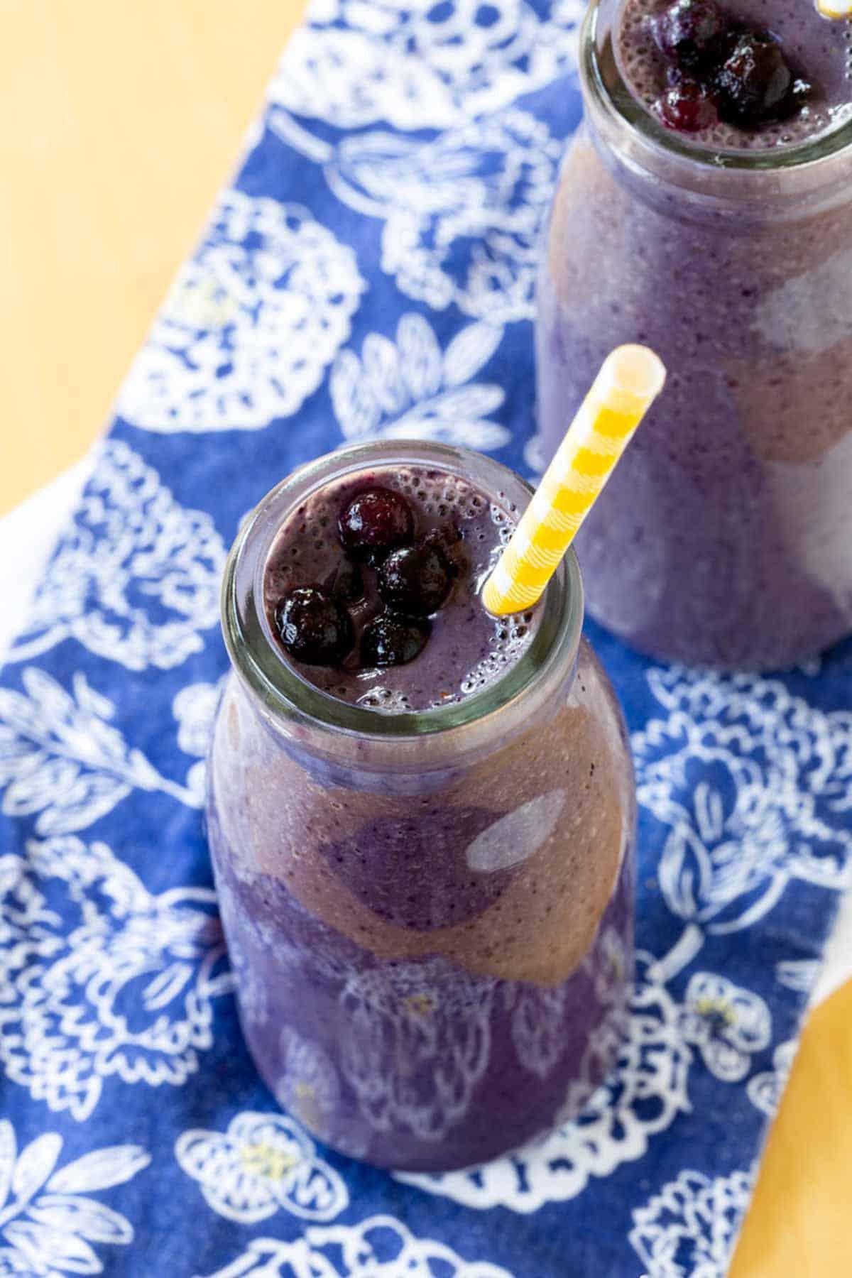 Two blueberry smoothies in glass bottles on top of a blue cloth napkins with white flowers.
