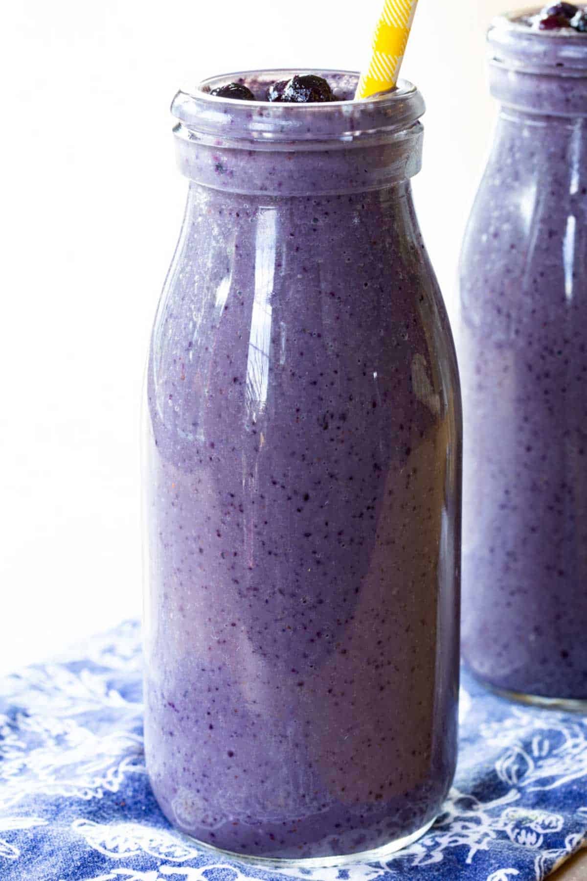 A tall glass bottle filled with a blueberry smoothie on a blue cloth napkin with a white design.