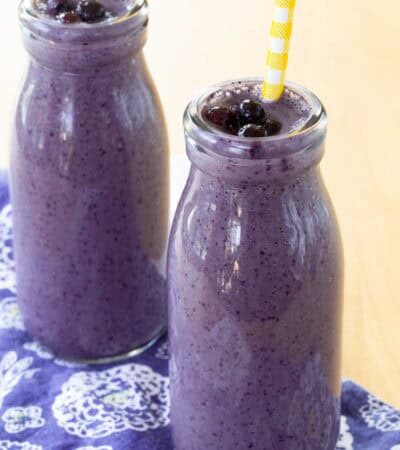 Two Blueberry Smoothies in glass bottles garnished with small thawed wild blueberries.