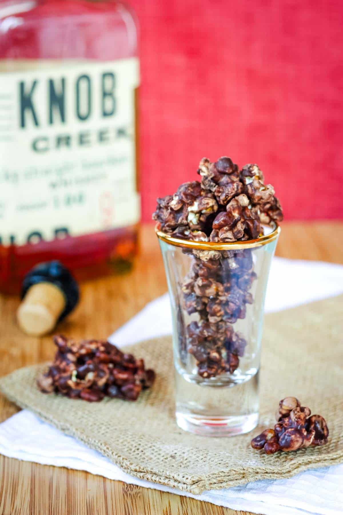 A shot glass filled with popcorn and pecans covered in chocolate with a bottle of Knob Creek bourbon in the background.