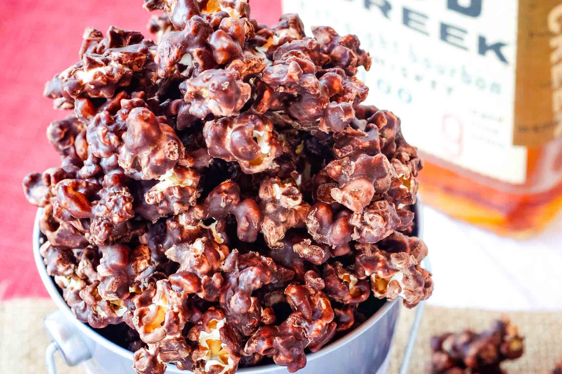 Chocolate popcorn in a bucket with a bottle of bourbon in the background.