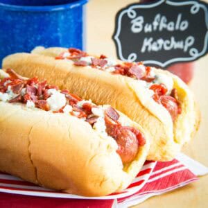 Two Buffalo Hot Dogs topped with with Bacon and Blue Cheese in buns on a red and white striped plate.