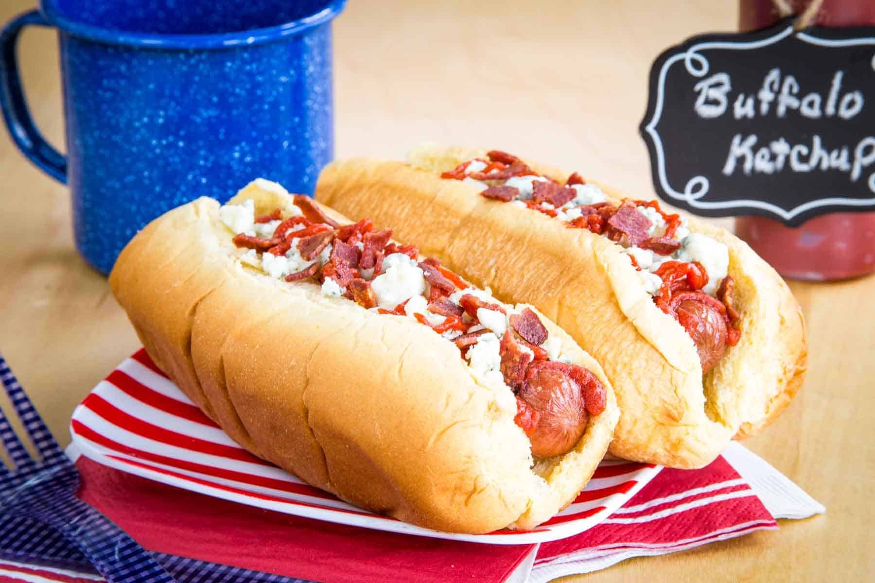 Two hot dogs in buns topped with buffalo ketchup, bacon, and crumbled blue cheese on a striped plate.