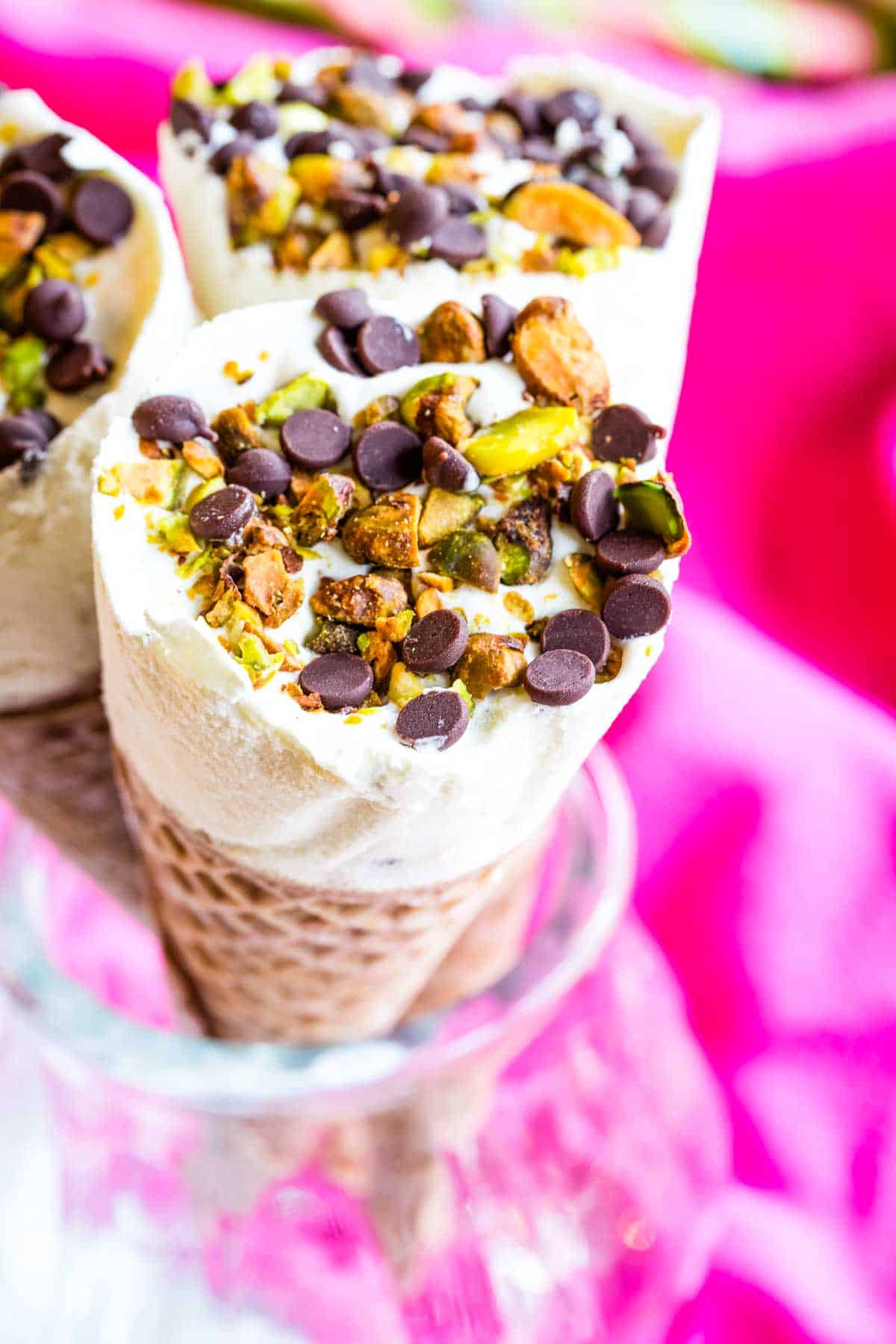A closeup of an ice cream cone topped with mini chocolate chips and chopped pistachios.