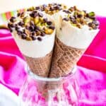 Three ice cream cones topped with mini chocolate chips and pistachios in a glass jar with text overlay that says "Cannoli Ice Cream Cones".