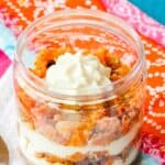 A jar of a carrot cake trifle with layers of crumbled cake and cream cheese filling on top of colorful cloth napkins.