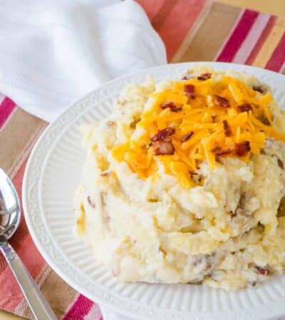 A bowl of mashed potatoes with bacon and cheddar cheese on a striped placemat with a serving spoon next to it.