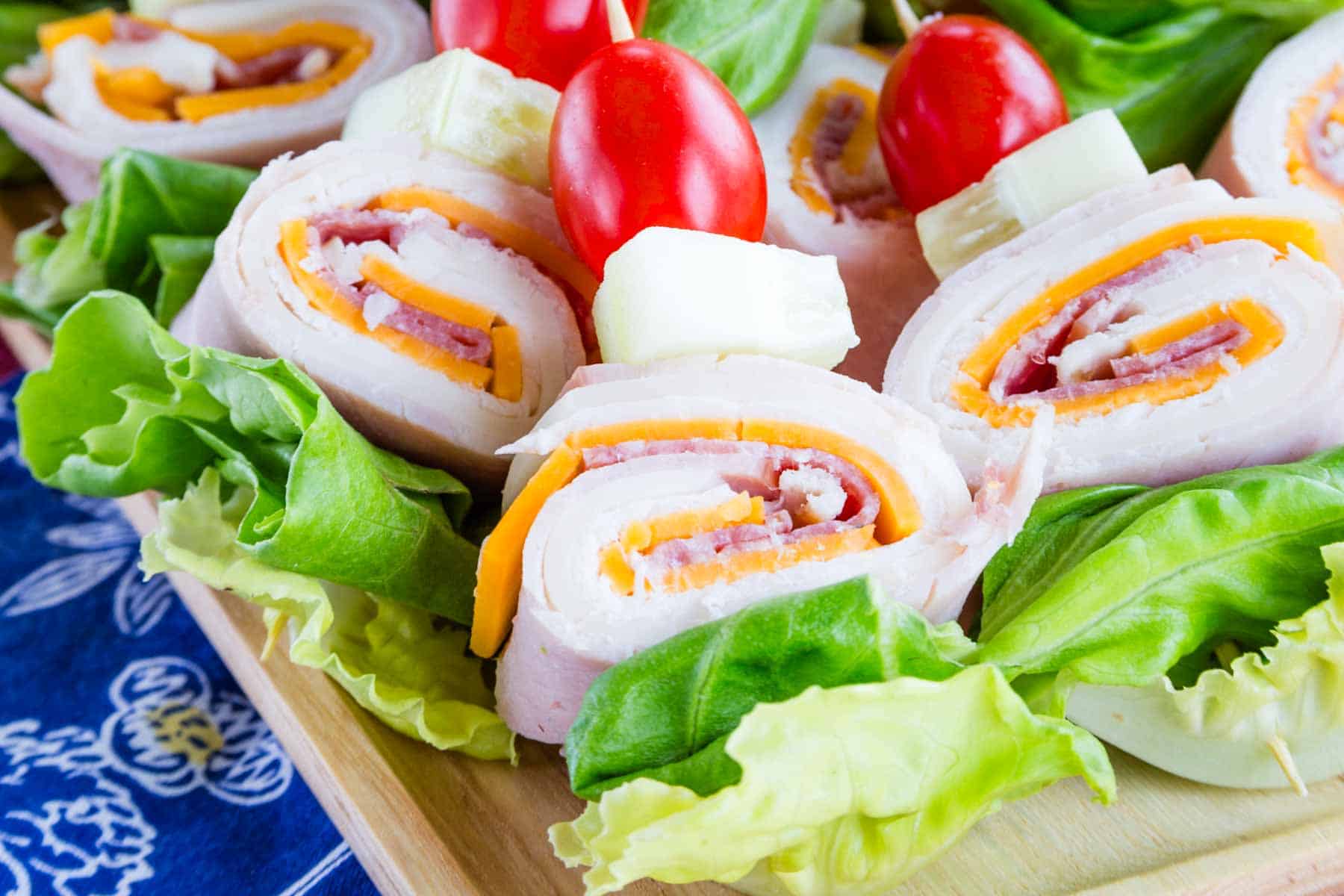 Rolls of deli meats and cheese threaded onto toothpick with lettuce, cucumber and tomato on a wooden platter.