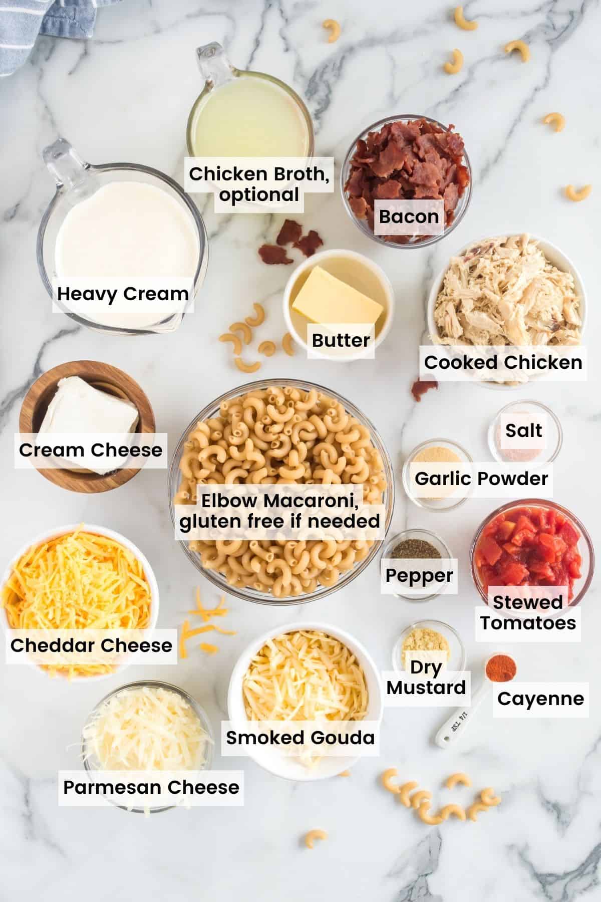 Bowls and measuring cups and spoons holding elbow noodles, chicken broth, heavy cream, cream cheese, cheeses, spices, chicken, bacon, and tomatoes withg text labels.