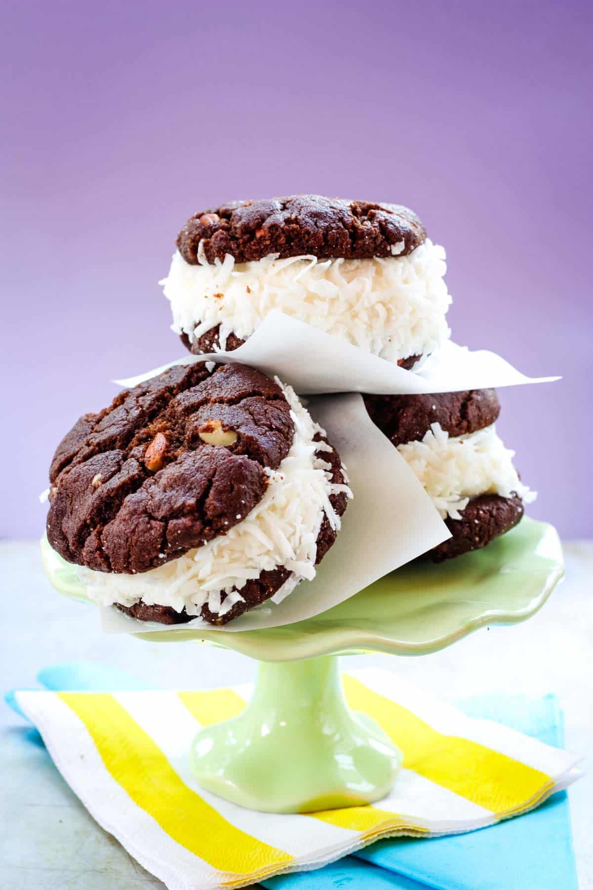 Three ice cream sandwiches made with coconut ice cream and two chocolate almond cookies leaning against each other on a small green cake stand.