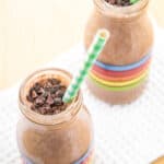 Looking down at two glass bottles filled with a chocolate smoothie with a straw and cacao nibs on top.