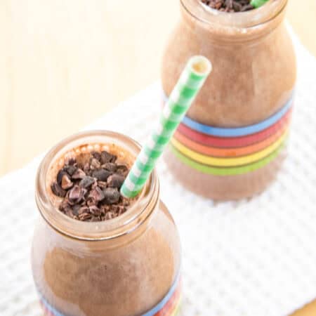 Looking down at two glass bottles filled with a chocolate smoothie with a straw and cacao nibs on top.