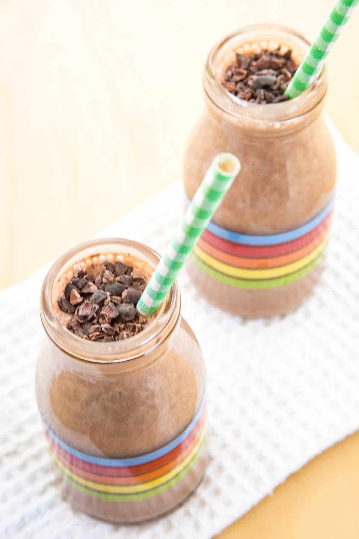 Looking down at two glass bottles filled with a chocolate smoothie with a straw and cacao nibs on top.