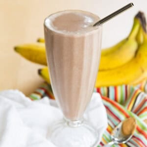 A chocolate peanut butter smoothie in a tall milkshake glass with bananas in the background.