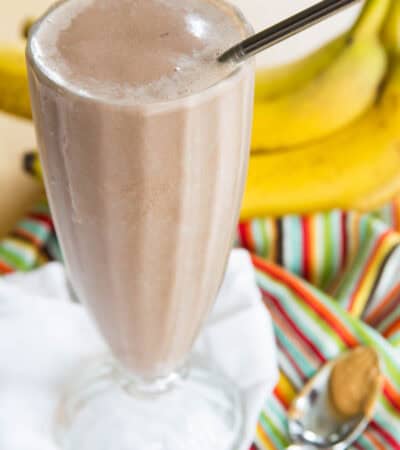 A Chocolate Peanut Butter Banana Smoothie in a tall glass with a spoon of peanut butter next to it.