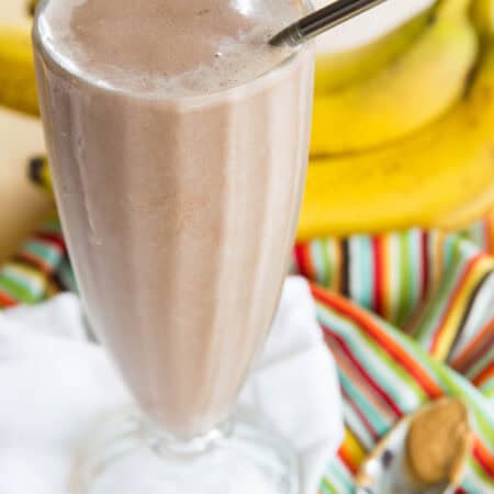 A Chocolate Peanut Butter Banana Smoothie in a tall glass with a spoon of peanut butter next to it.