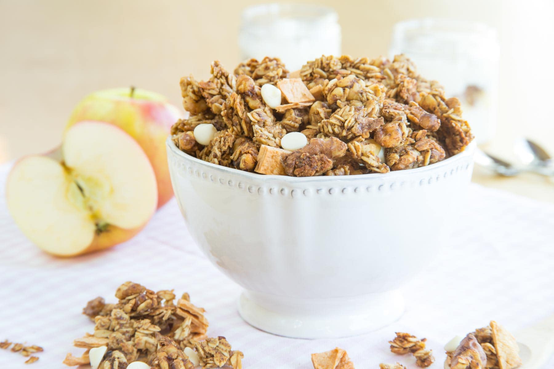 A bowl of granola surrounded by some of the clusters on the table and apples in the background.