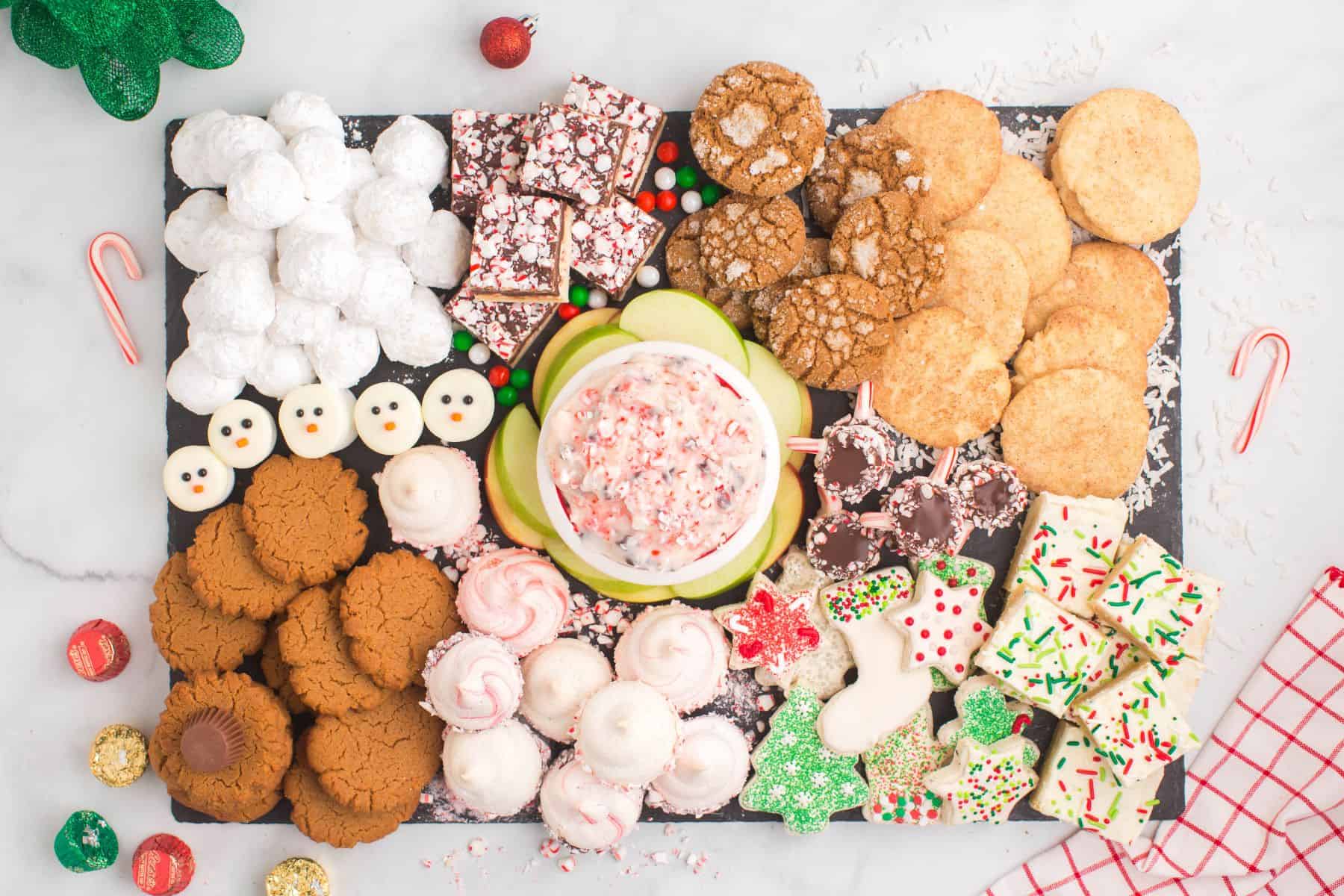 A cookie dessert board with an assortment of cookies, candies, and apple slices and a bowl of peppermint dip on a slate platter.